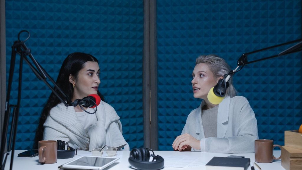 Two women engaged in conversation, recording podcast in studio with microphones and headphones.