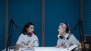 Two women in a soundproof studio recording a podcast with microphones and equipment.