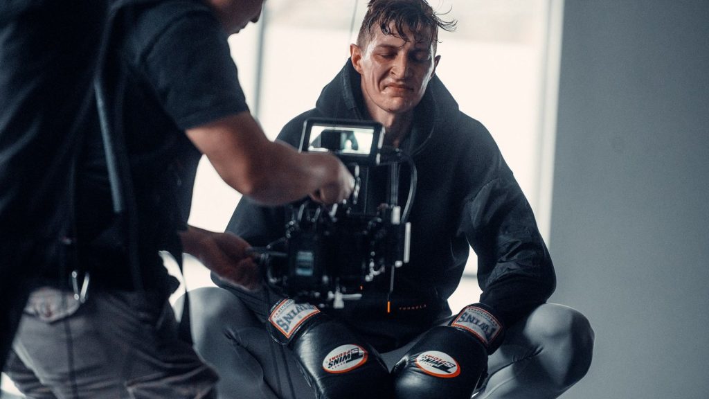 A boxer crouches in studio, with a camera crew capturing the moment, showcasing determination and athleticism.