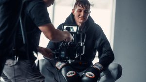 A boxer crouches in studio, with a camera crew capturing the moment, showcasing determination and athleticism.