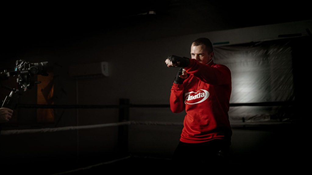 Powerful male athlete in sportswear practicing boxing hits in ring against crop anonymous partner recording video on pho