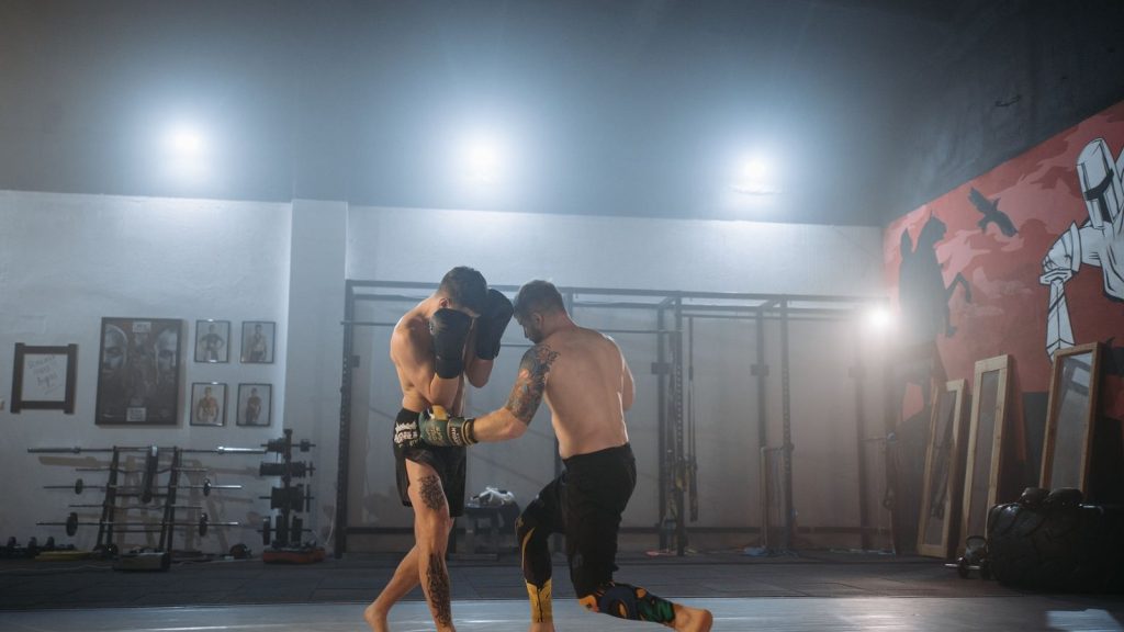 Two men engaged in an intense MMA training session inside a gym under dramatic lighting.