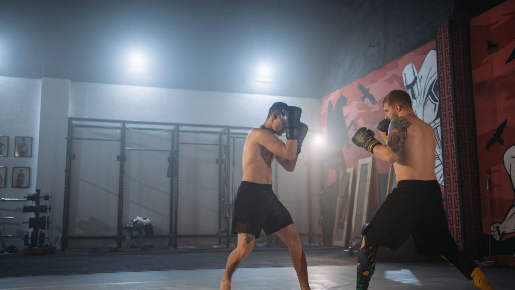 Two muscular men sparring in an MMA gym with gloves, showcasing fitness and combat skills.