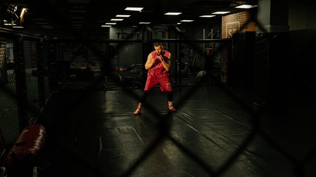 Male athlete wearing red activewear training intensely in an indoor martial arts gym. Dark and focused setting.