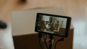A camera monitor shows a scene with a person sitting among boxes, highlighting the filming process.