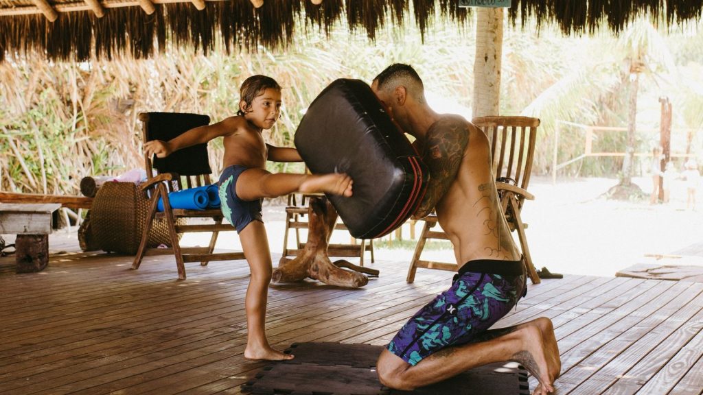 A father and son duo practice martial arts in a tropical outdoor setting, engaging in a training session.