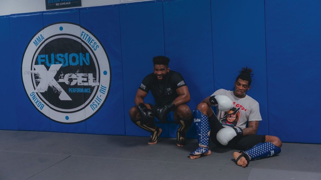 Two young men sitting and resting in an MMA gym, wearing boxing gloves and gear, smiling.