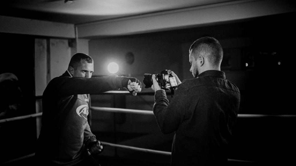 Black and white of determined male athlete demonstrating boxing hit against partner recording video on photo camera in r