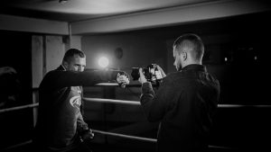 Black and white of determined male athlete demonstrating boxing hit against partner recording video on photo camera in r
