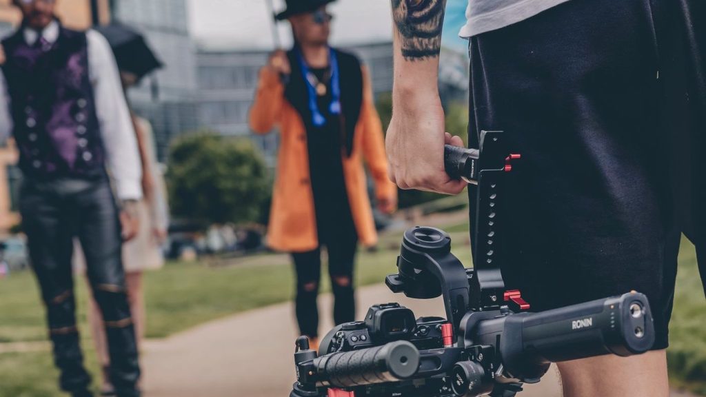 A tattooed videographer films people in cosplay costumes with a gimbal outdoors.