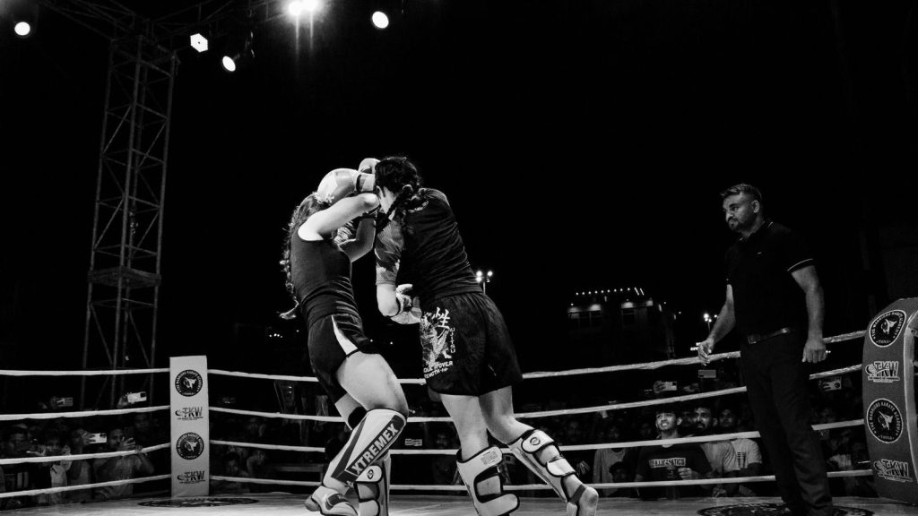 Two female fighters in an intense boxing match captured in dramatic black and white photography.