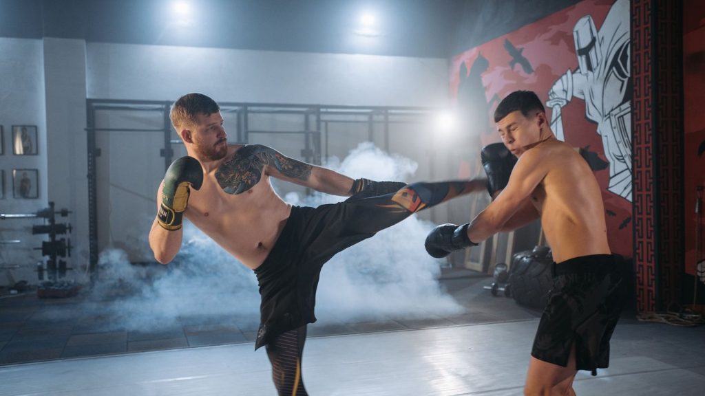 Two shirtless men engaging in vigorous MMA sparring, demonstrating skill and intensity indoors.