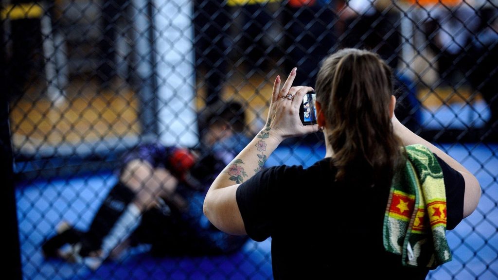 A woman records a mixed martial arts match using her smartphone inside the ring.