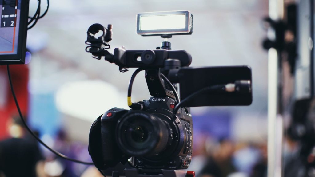A close-up shot of a Canon video camera with attached equipment in a blurred indoor setting.