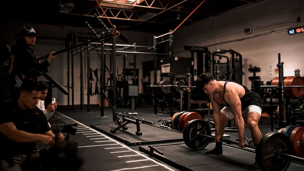 Muscular man performs deadlift in a gym with film crew documenting, showcasing strength and focus.
