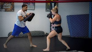 A man and woman engage in mixed martial arts training inside an indoor gym.
