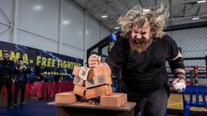 A strong man demonstrates brick breaking skills in an indoor MMA competition setting.
