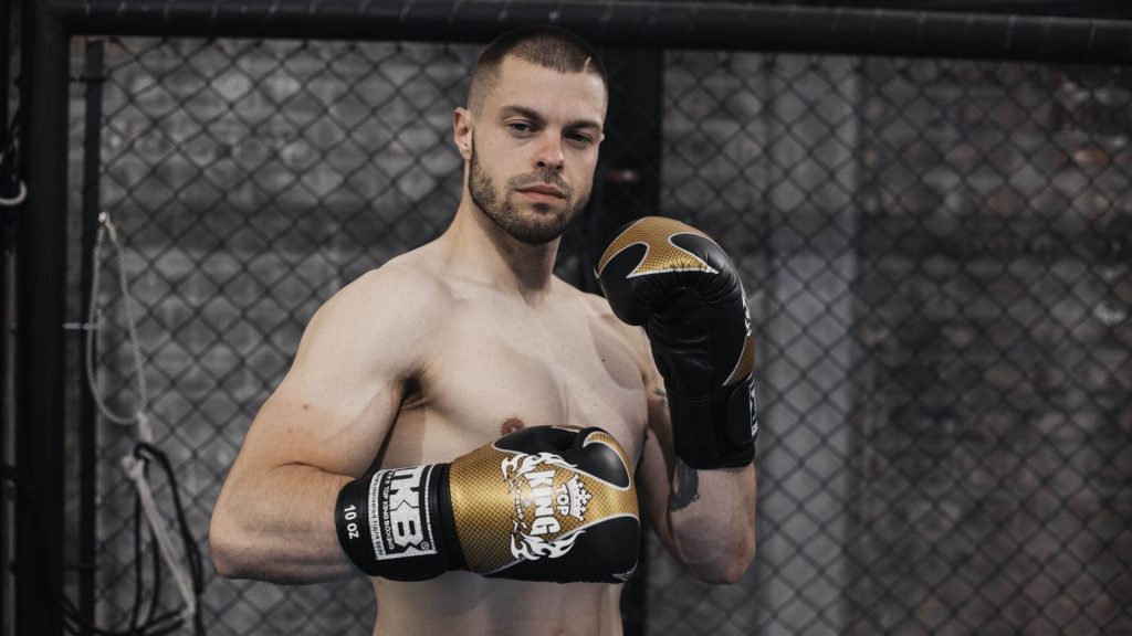 Shirtless male boxer with gloves posing in an indoor training arena, showcasing strength and intensity.