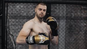 Shirtless male boxer with gloves posing in an indoor training arena, showcasing strength and intensity.