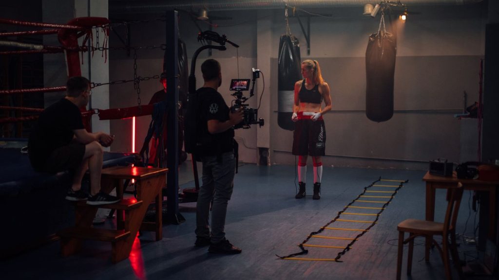 Female boxer preparing in a dimly lit boxing gym with a film crew.