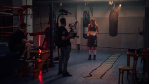 Female boxer preparing in a dimly lit boxing gym with a film crew.